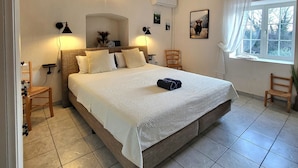 A neatly arranged bedroom with a large bed, two bedside tables, a chair, a window with curtains, and a wall-mounted air conditioner.