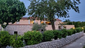 A stone-walled building with a tiled roof, surrounded by lush greenery and a paved pathway.