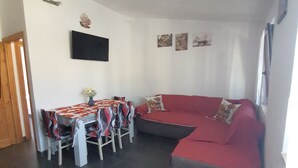 A living room with a red sofa, a dining table with chairs, a flat-screen TV mounted on the wall, and a small coffee machine. - Ana&julio holiday home (Valledoria)
