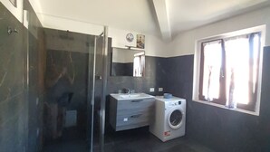A compact bathroom with a glass shower enclosure, a built-in sink, a washing machine, and a window with curtains. - Ana&julio holiday home (Valledoria)