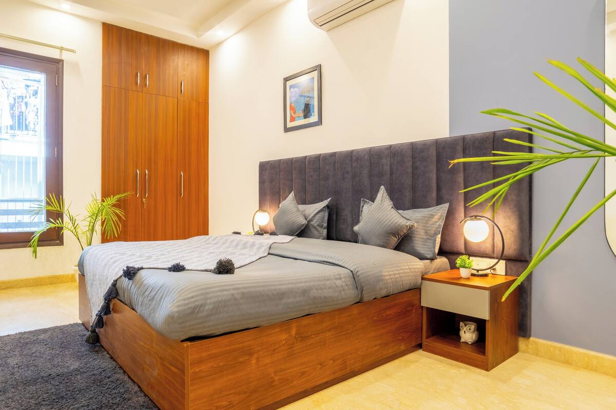 Une chambre moderne avec un lit en bois, une literie grise, une tête de lit grise, des lampes de chevet, une petite table de chevet, une plante en pot et un tableau encadré au mur.