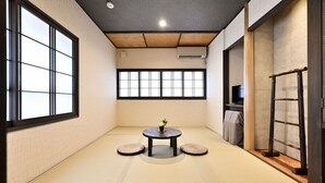 A tatami mat room with a tatami floor, a round table with a vase, and a TV on the wall. - Wooribow Ryokan (Kusatsu)