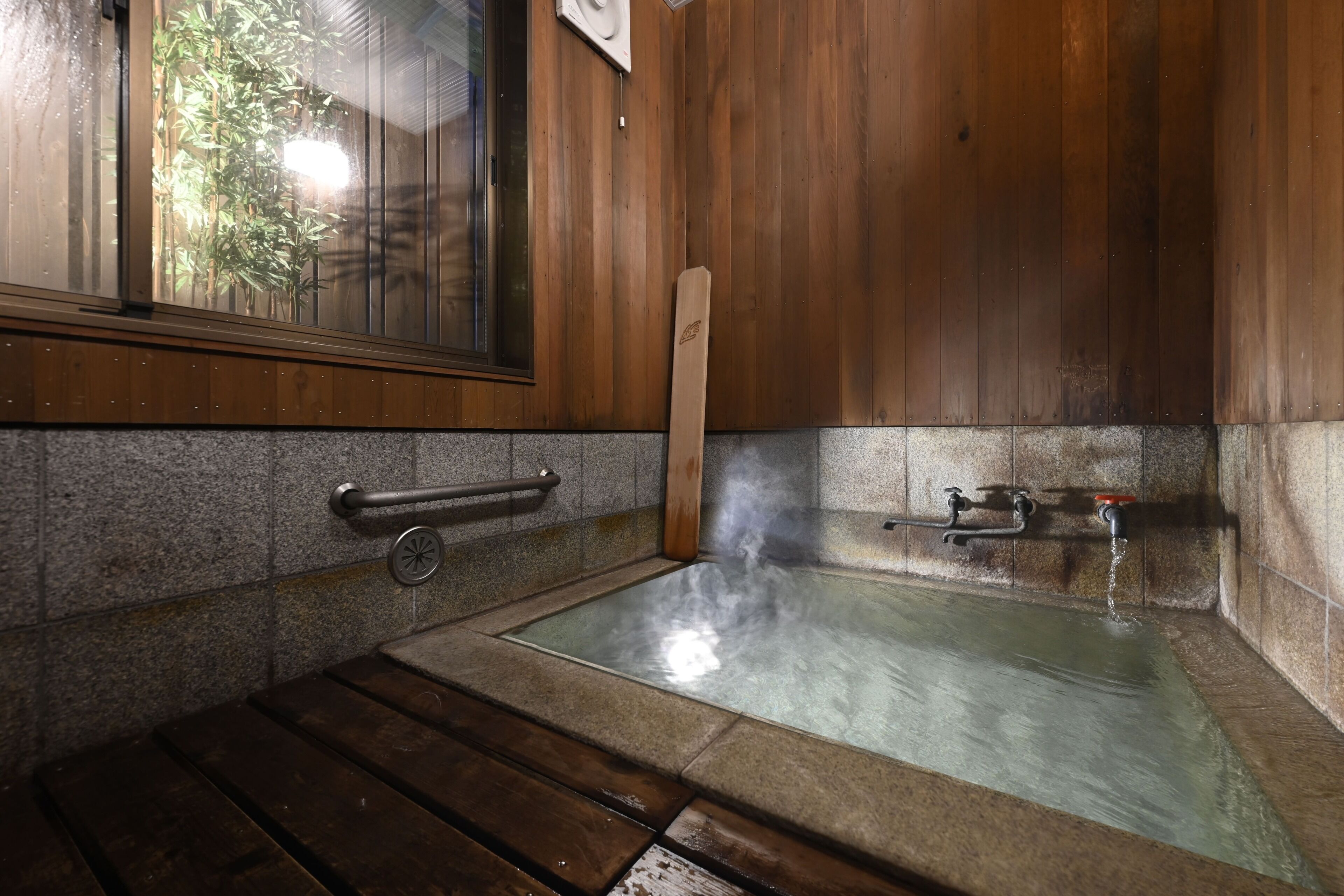 Une pièce en bois avec un bain alimenté par une source d’eau chaude, une fenêtre donnant sur un jardin de bambous et un mur en pierre.