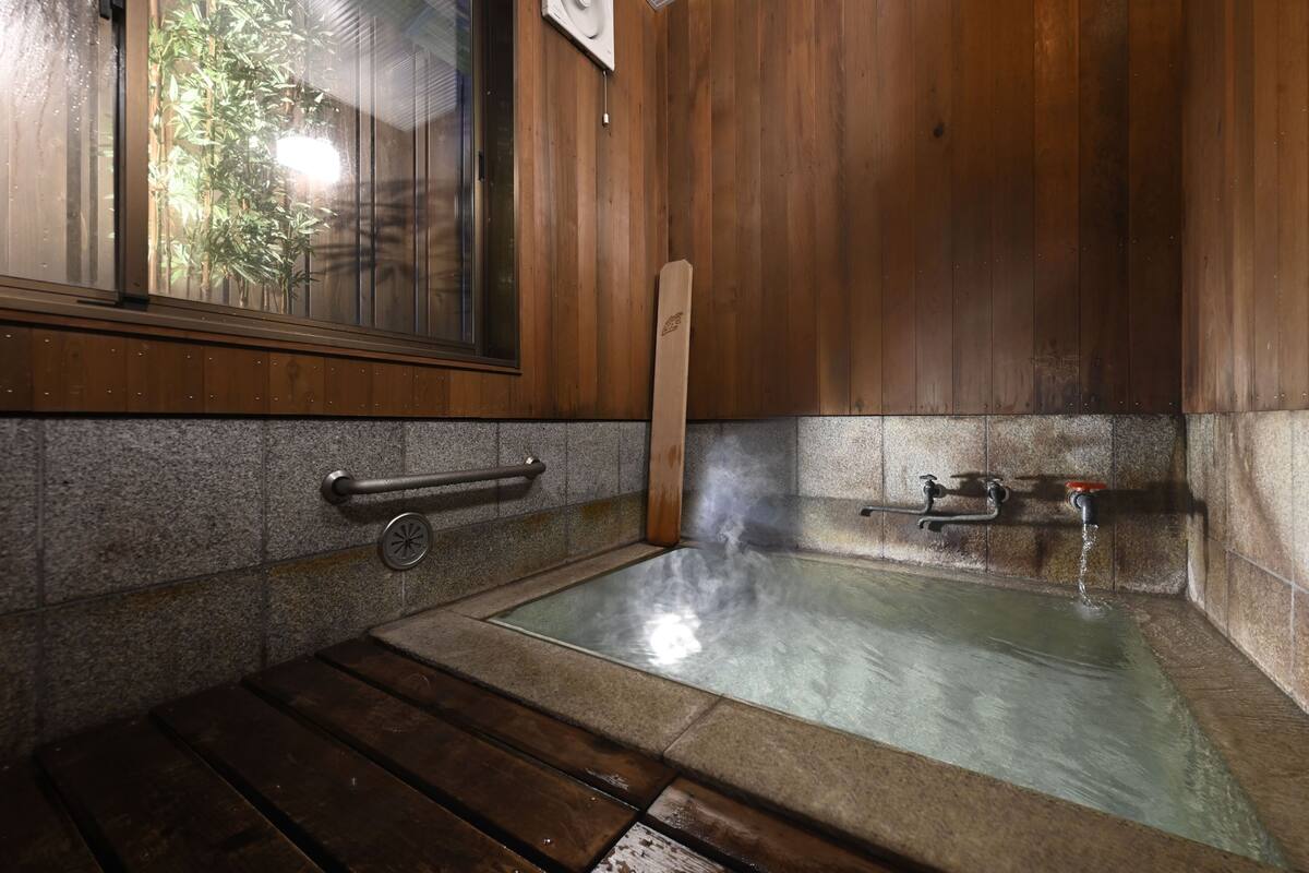 Una habitación de madera con un baño de aguas termales, una ventana con bambú en el exterior y una pared de piedra.