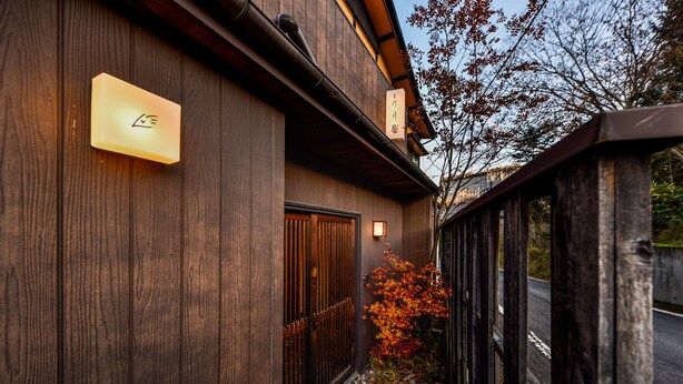 A wooden building with a signboard, a gate, and a tree with autumn leaves. - Wooribow Ryokan (Kusatsu)