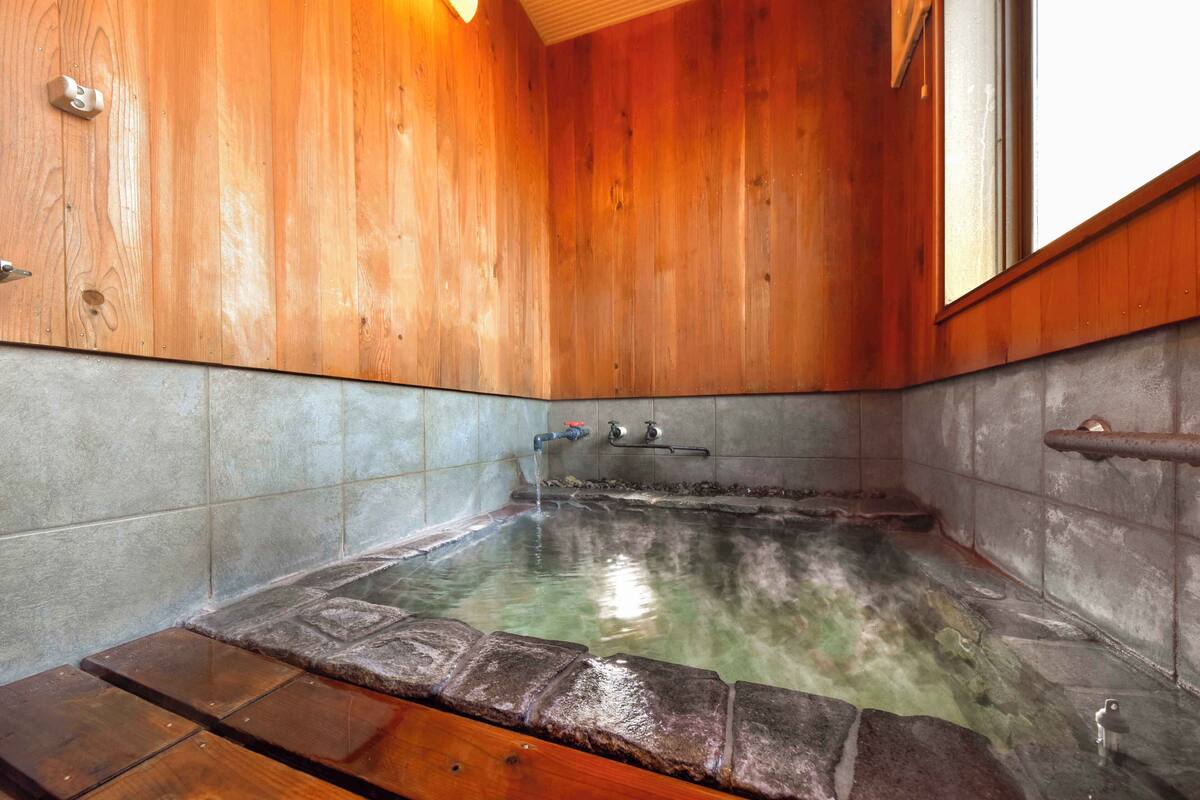 Interior de madera con una bañera caliente de baldosas de piedra, un banco de madera y una ventana.
