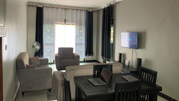 Een woonkamer met een bank, fauteuil en een eettafel met stoelen. Er is een televisie aan de muur en een ventilator op de vloer.