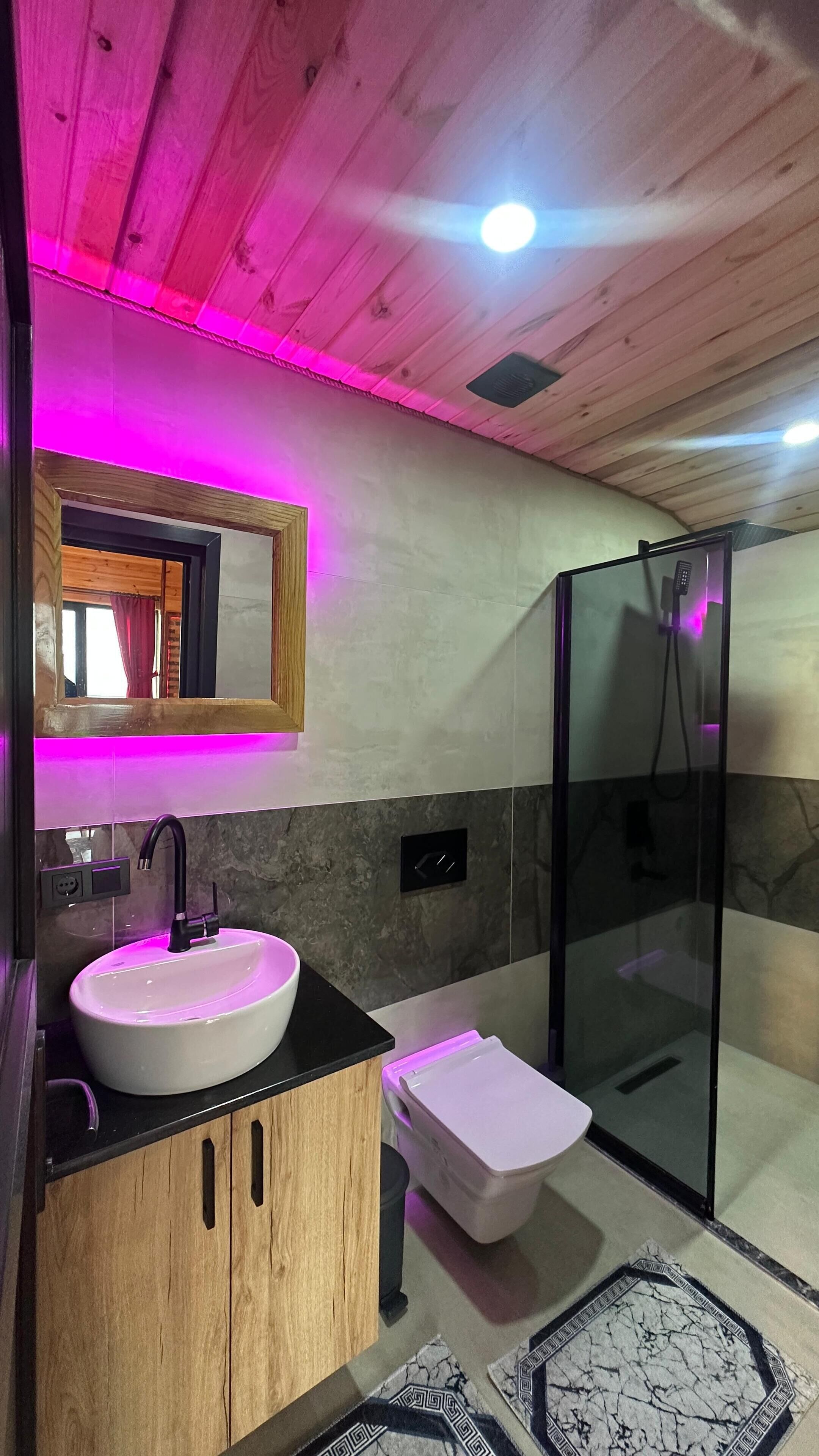 A modern bathroom with a wooden vanity, a white sink, a glass shower enclosure, and a marble-patterned floor.