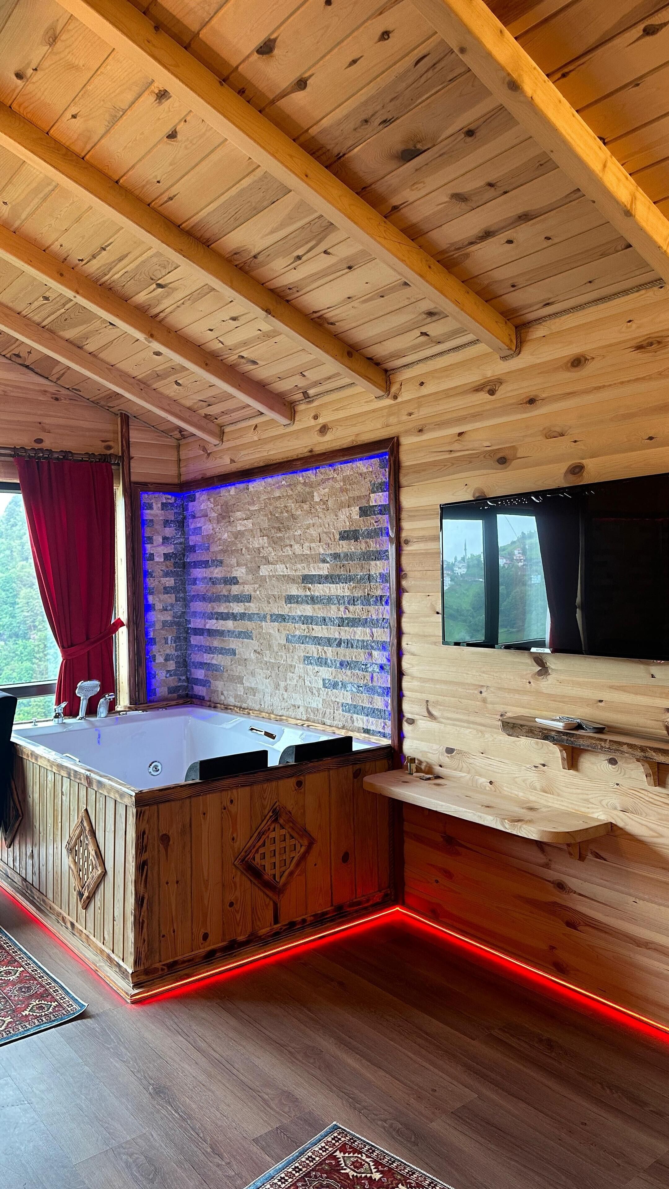 A wooden cabin interior with a wooden ceiling, a brick wall with a blue light strip, a wooden bar, and a large window.