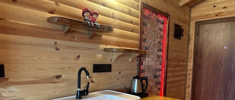 Interior de una cabaña de madera con una cocina que incluye un fregadero, un hervidor y una repisa con figuritas.