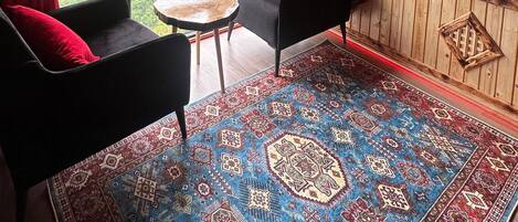 Una habitación con vigas de madera en el techo, un ventanal con cortinas rojas, un sofá negro con un cojín rojo, una mesita redonda pequeña y una alfombra estampada.