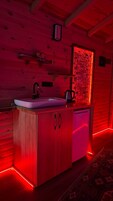 Interior de una cabaña de madera con un lavabo iluminado en rojo, armarios y un refrigerador pequeño.