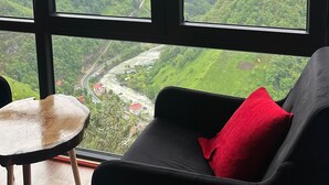 Een kamer met een groot raam dat uitzicht geeft op een groene vallei en een rivier. Er staat een zwarte stoel met een rood kussen en een klein rond tafeltje met een stoel.