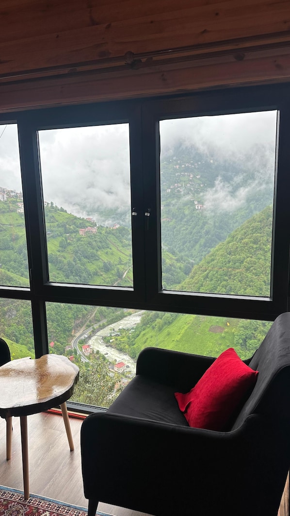 Habitación con un ventanal grande que ofrece vistas a un valle verde y un río. Hay una silla negra con un cojín rojo y una pequeña mesa redonda con una silla.
