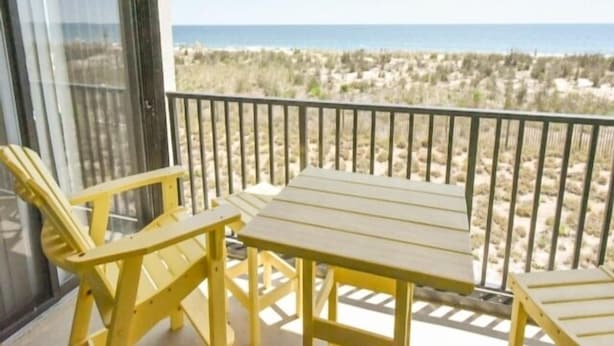 Ein Balkon mit Tisch und zwei Stühlen, mit Blick auf einen Strand mit Dünen.