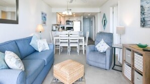 Ein modernes Wohnzimmer mit einem blauen Sofa, einem grauen Sessel und einem Essbereich mit weißem Tisch und Stühlen.