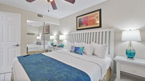 Une chambre d’hôtel avec un grand lit, deux tables de chevet, un ventilateur de plafond, un tableau encadré et un miroir.