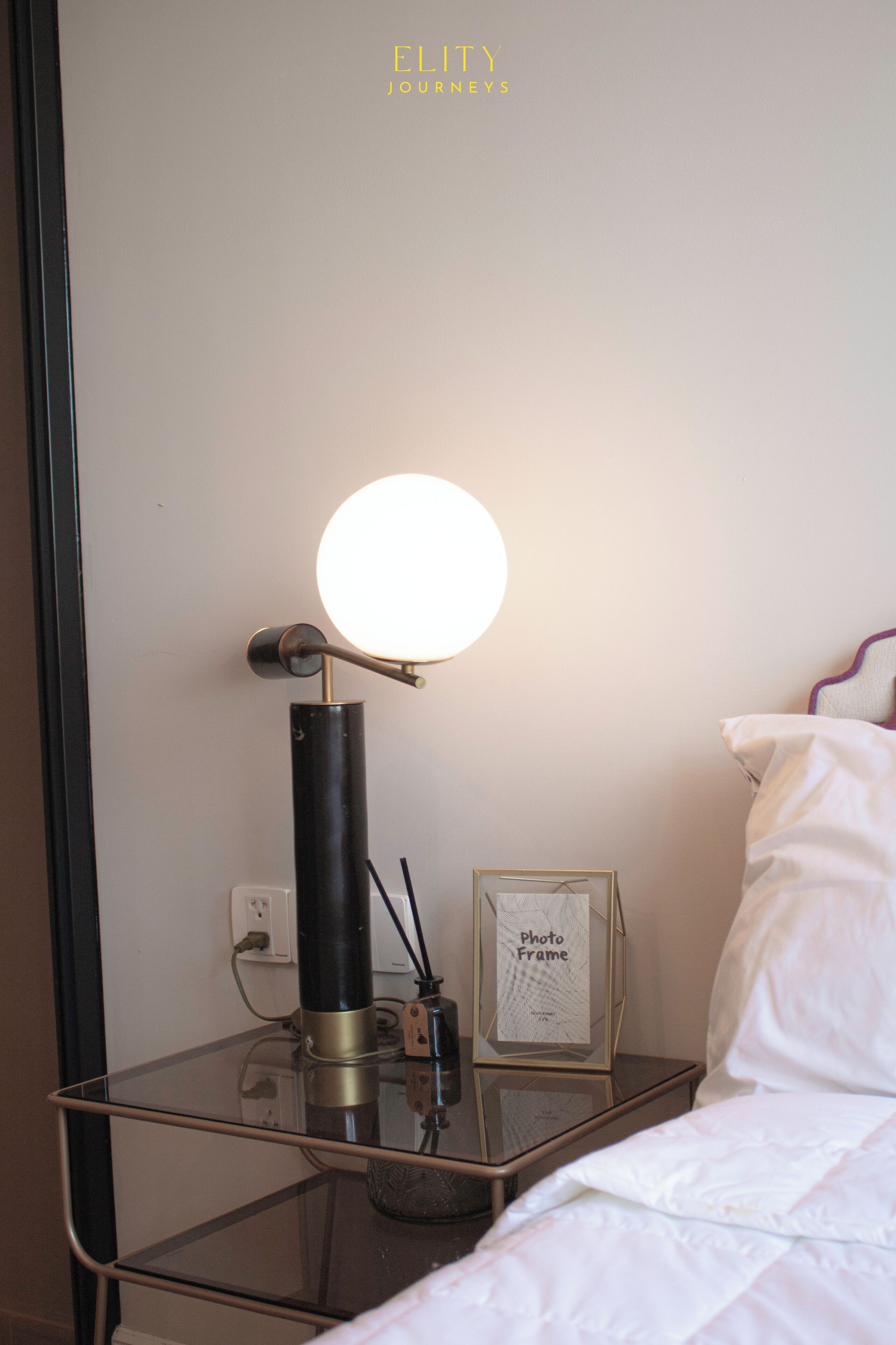 A neatly made bed with white bedding, a bedside table with a glass top, a black lamp with a round light, and a framed photo frame on the table.