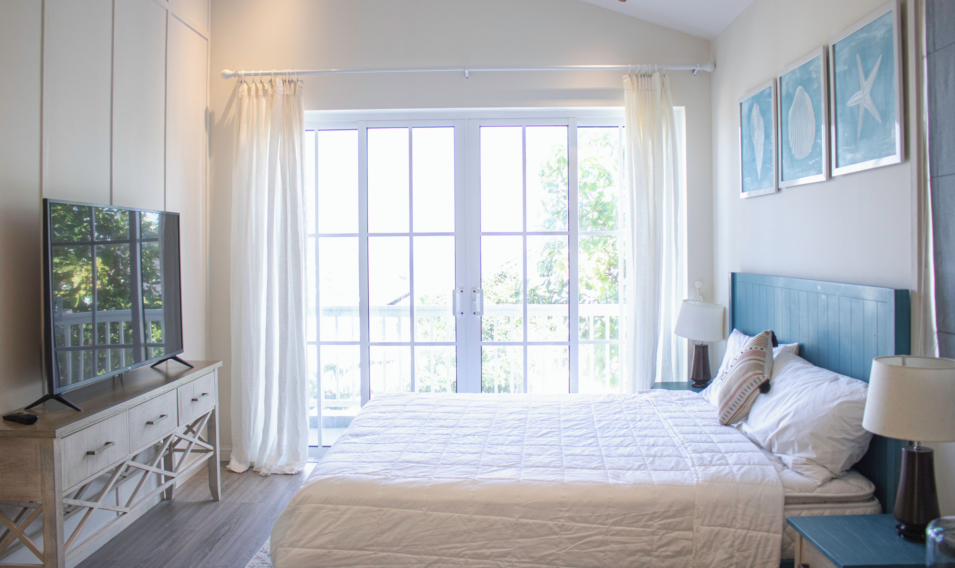A bedroom with a large bed, a TV on a wooden dresser, and a view of the outdoors through a large window.