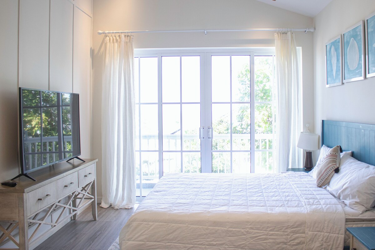 A bedroom with a large bed, a TV on a wooden dresser, and a view of the outdoors through a large window.