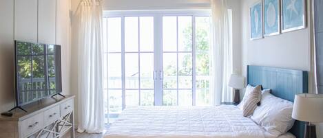 A bedroom with a large bed, a TV on a wooden dresser, and a view of the outdoors through a large window.