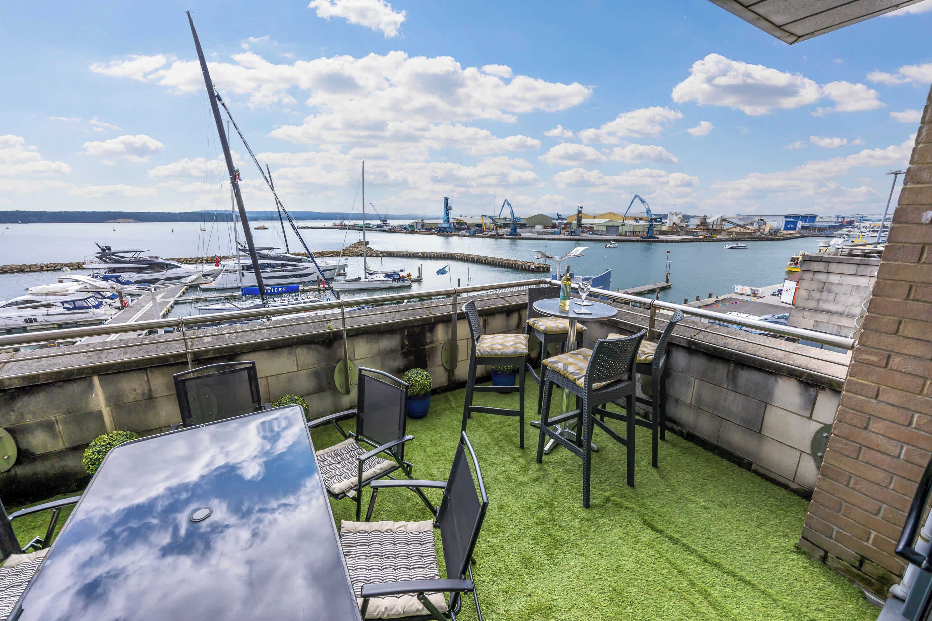 Une terrasse avec des chaises et une table, donnant sur une marina où sont amarrés des bateaux et où se trouvent des structures industrielles.