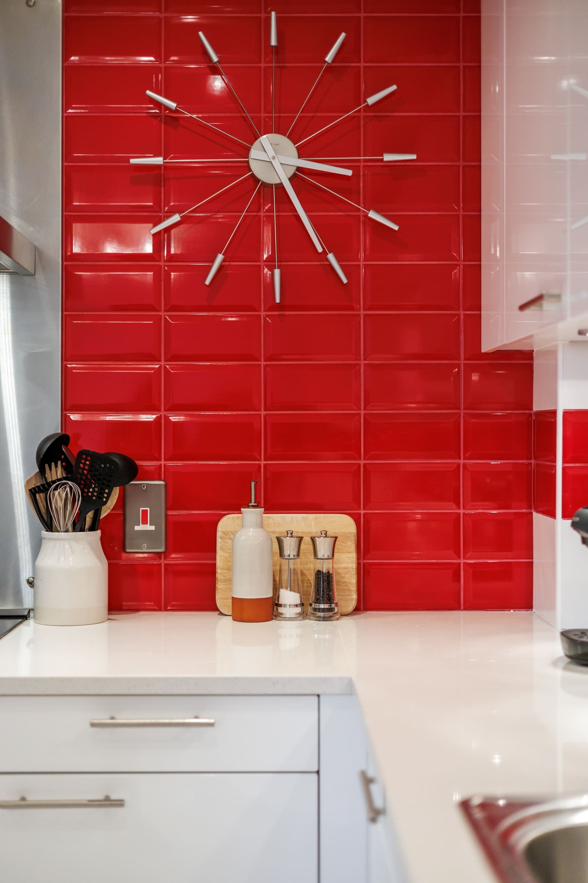 Une cuisine avec un mur recouvert de carreaux rouges, une horloge blanche et divers ustensiles de cuisine posés sur le plan de travail.