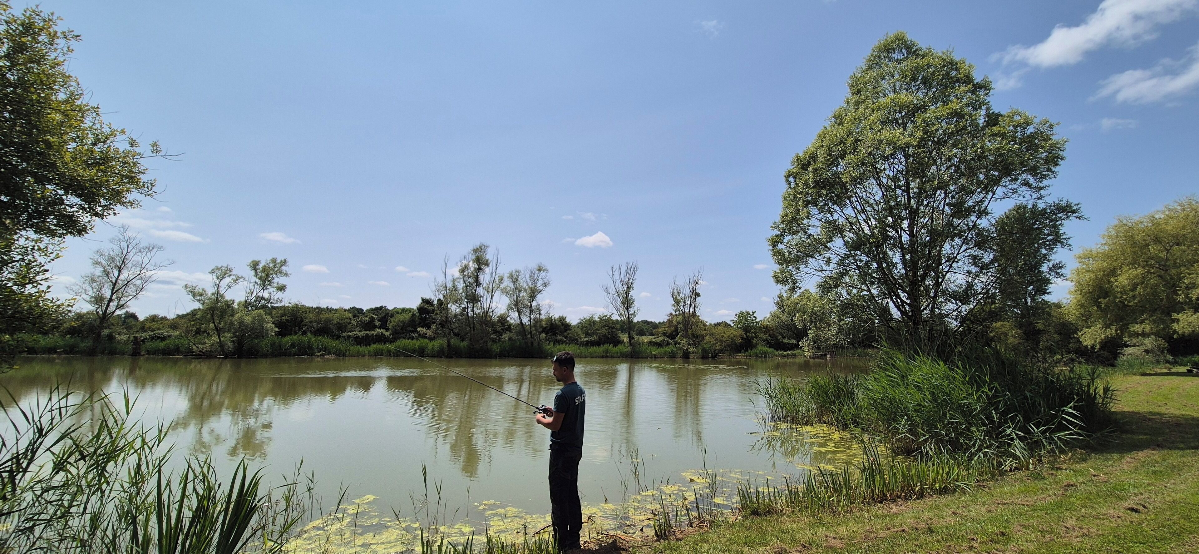 Une personne est en train de pêcher au bord d’un lac paisible, entouré d’arbres et de verdure.