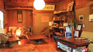 Ein traditioneller japanischer Raum mit Holzwänden, Tatami-Bodenbelag, einem niedrigen Tisch, Regalen mit diversen Gegenständen und einer hängenden Leuchte.