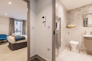 Ein modernes Hotelzimmer mit einem Bett, einem blauen Sessel, einem Badezimmer mit Toilette, Waschbecken und Spiegel.