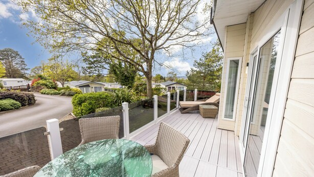 A covered outdoor area with wicker furniture and a glass table, overlooking a road and greenery.