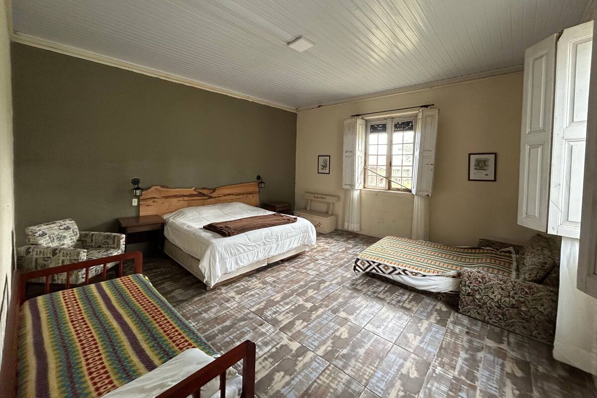 Un dormitorio con una cama grande, una cama más pequeña, una silla, un banco y una ventana con contraventanas.