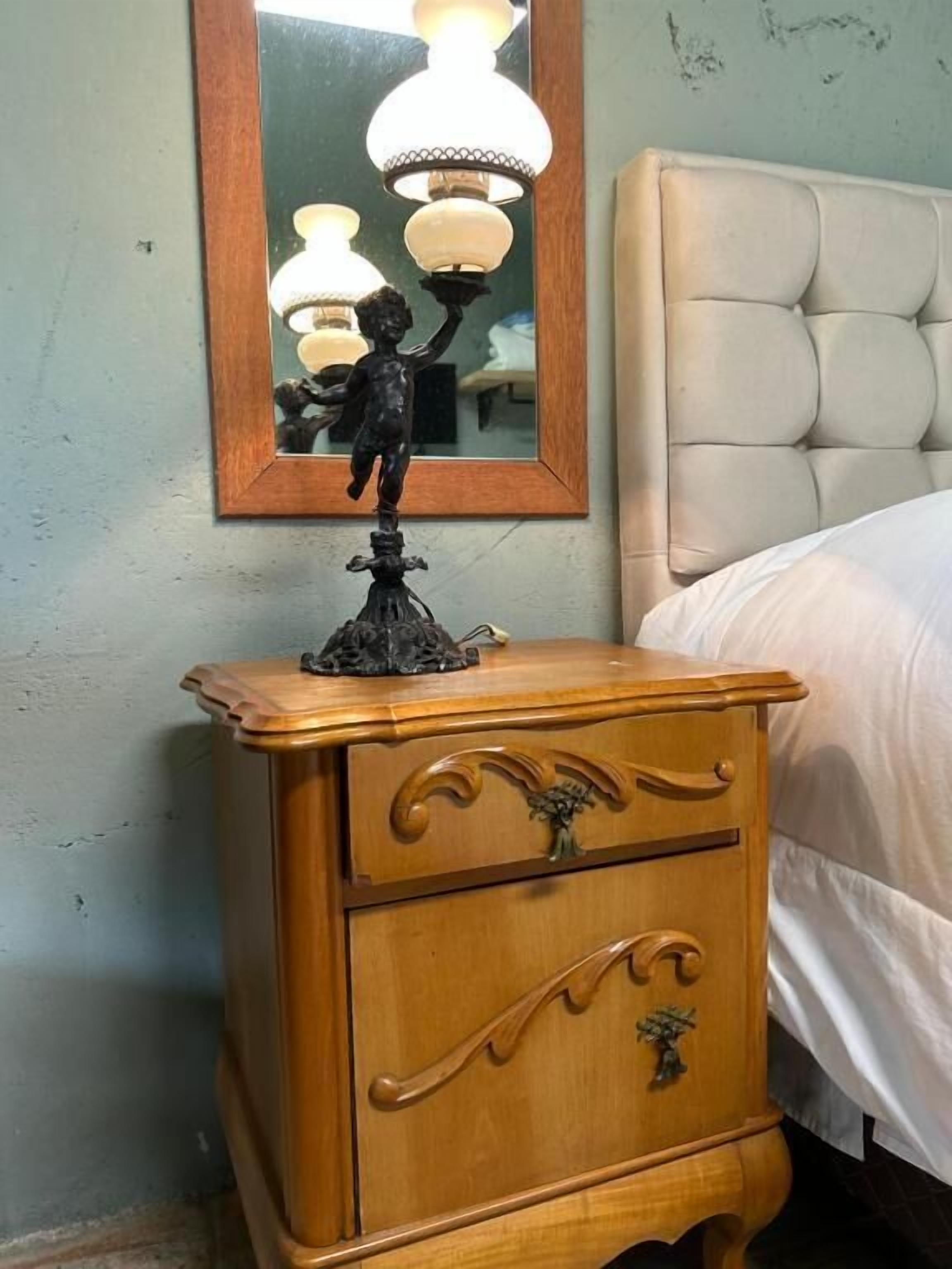 Uma mesa de cabeceira de madeira com uma luminária e uma estátua, um espelho, e uma cama com cabeceira.
