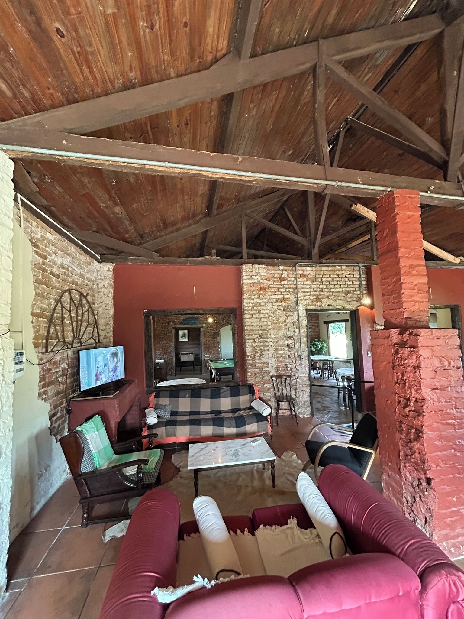 A rustic interior with exposed wooden beams, a brick wall, a checkered sofa, a television, and a billiards table.