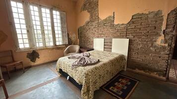 Une pièce avec un lit, une chaise et une fenêtre. Les murs sont endommagés et la brique est apparente.