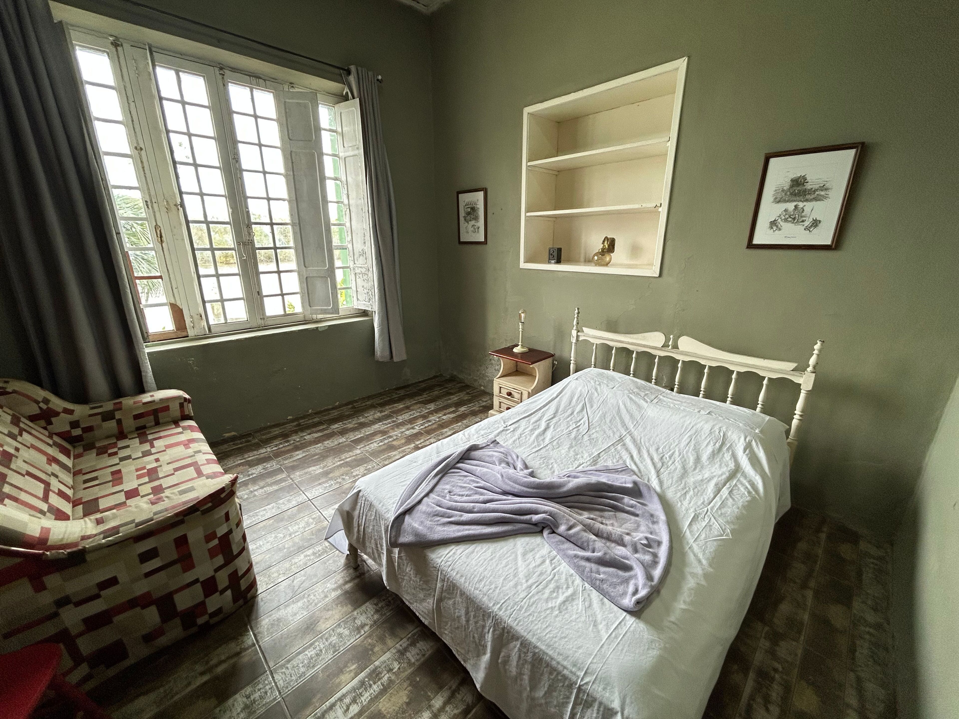 A bedroom with a bed, a chair, a window with curtains, and a shelf with pictures.