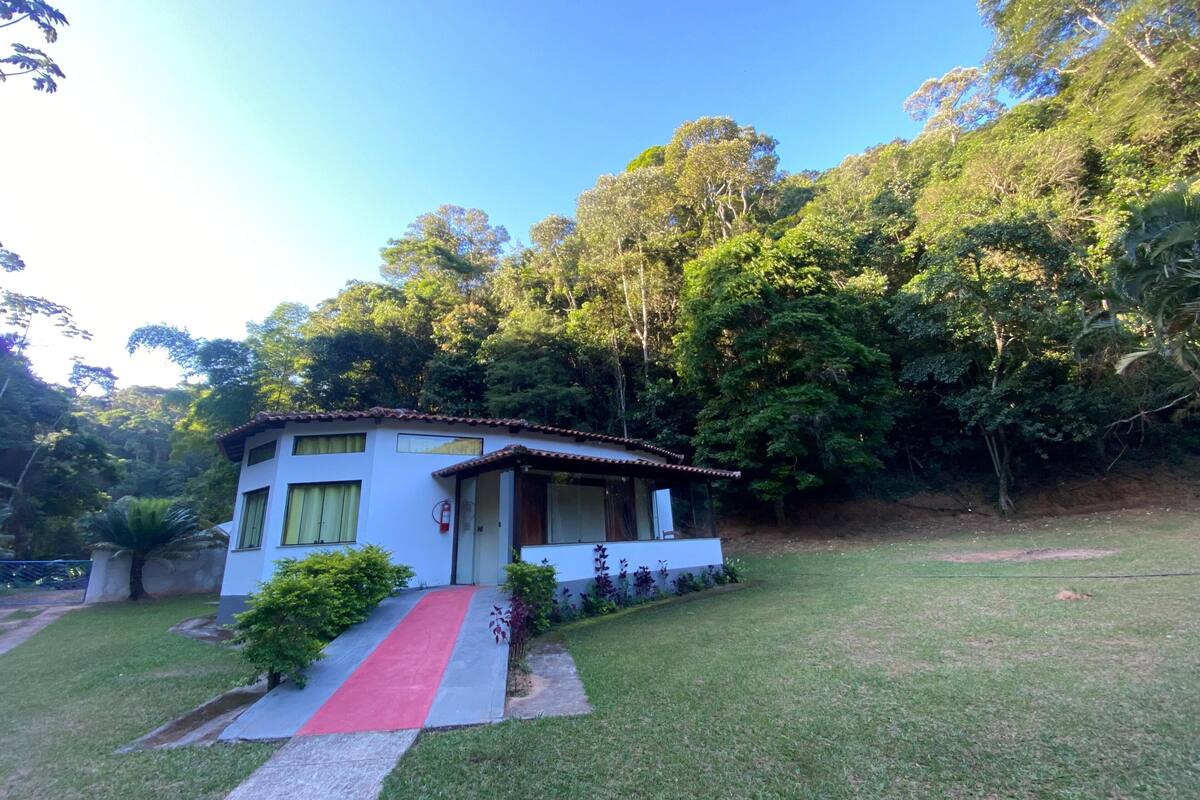 A small white house with a red pathway leading to the entrance, surrounded by lush greenery and trees.