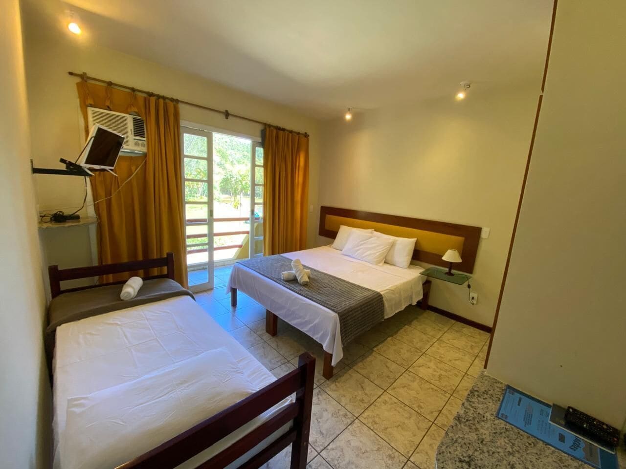 A hotel room with two single beds, a desk, a chair, a small table with a lamp, and an air conditioning unit.