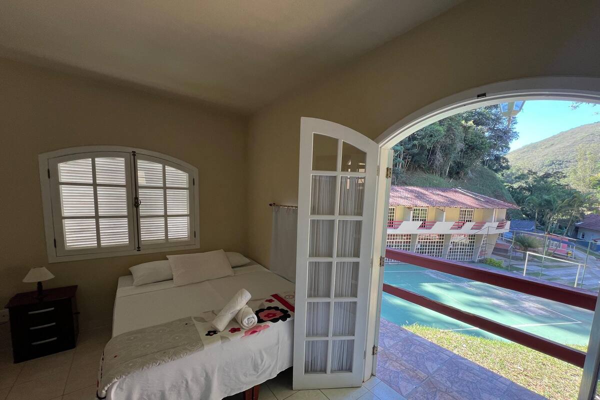A bedroom with a bed, a nightstand, a window with blinds, and an open door leading to a balcony with a view of a building and greenery.
