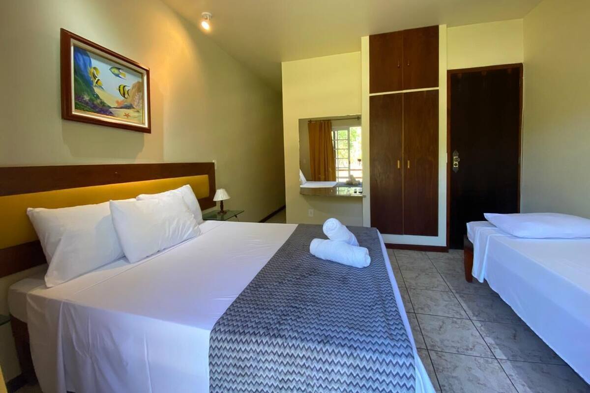 A hotel room with a large bed, two smaller beds, a wardrobe, a painting, and a bathroom visible through an open door.