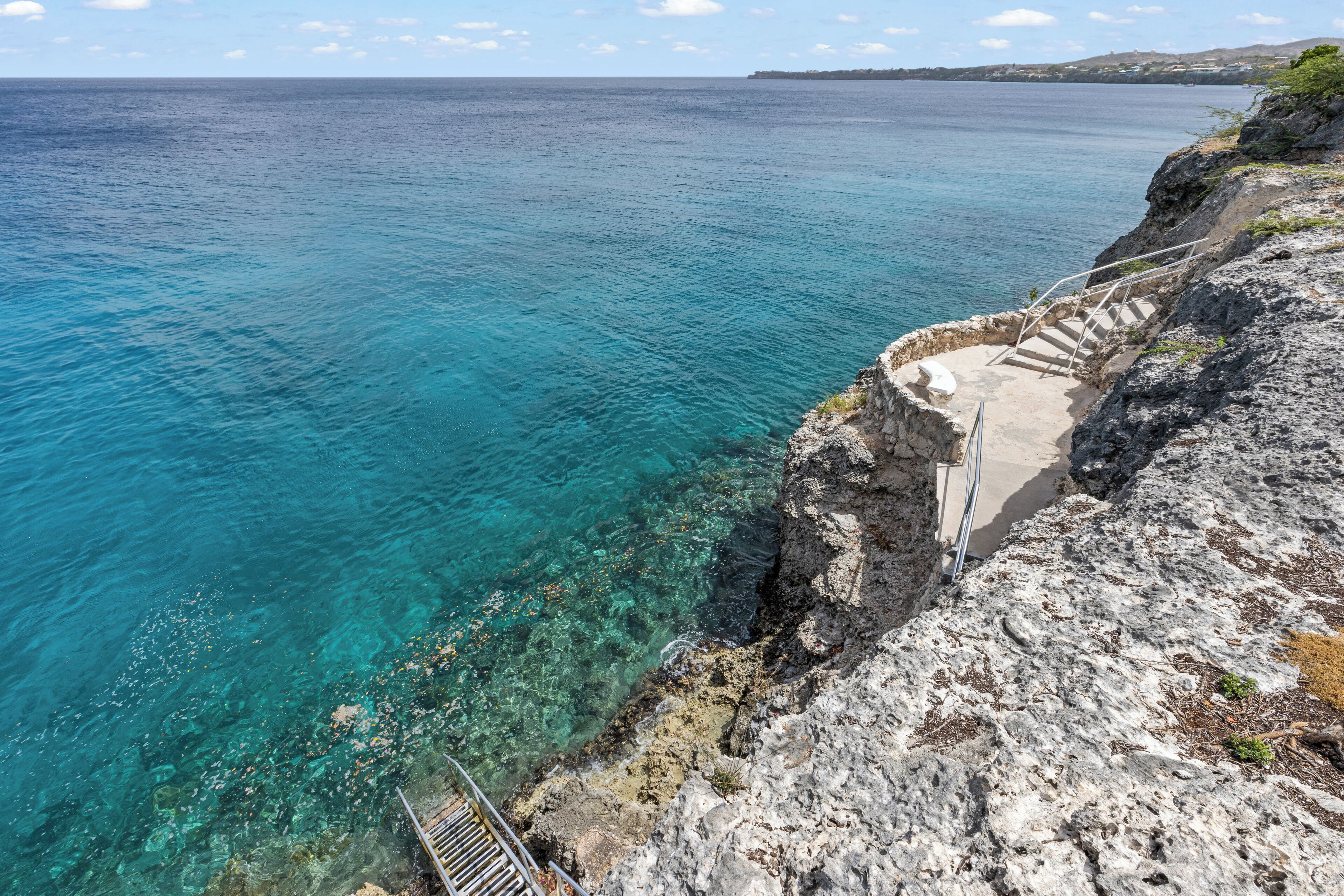 Une falaise côtière avec un escalier descendant vers la mer, une eau d’un bleu limpide et une côte visible au loin.