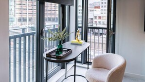 Un apartamento moderno con un ventanal grande, una mesita con un florero y libros, y un sillón cómodo.