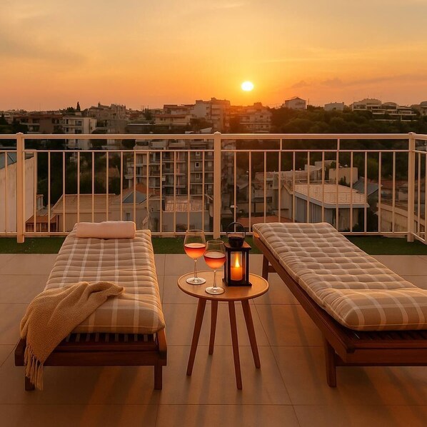 A balcony with two beds, a table with glasses and a lantern, and a sunset view.