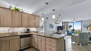 Een moderne keuken met houten kastjes, een vaatwasser en een granieten aanrecht. Er is een zithoek met een blauwe bank en een plafondventilator.