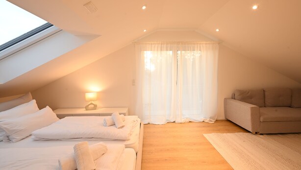 Un dormitorio con techo inclinado, una cama con ropa de cama blanca, un sofá, un ventanal con cortinas y suelo de madera.