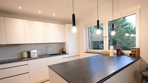 Una cocina moderna con armarios blancos, una isla grande, iluminación colgante y un ventanal con vistas a zonas verdes.