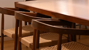 Una mesa de comedor de madera con sillas tejidas, lámparas colgantes y vista a una cocina.