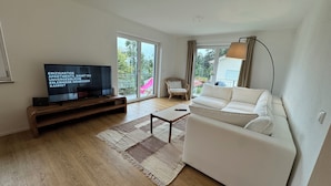 Una sala de estar moderna con un televisor de pantalla plana, un sofá blanco, una mesa de centro de madera y un ventanal con vistas al exterior.