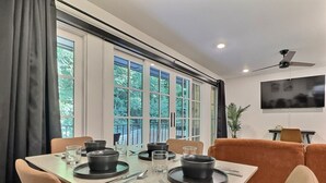 Uma área de refeições com uma mesa posta para quatro pessoas, grandes janelas com cortinas, um ventilador de teto e uma televisão na parede.