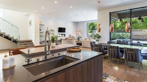 Cozinha moderna com ilha central, área de jantar com mesa e cadeiras grandes e sala de estar com sofá e televisão.